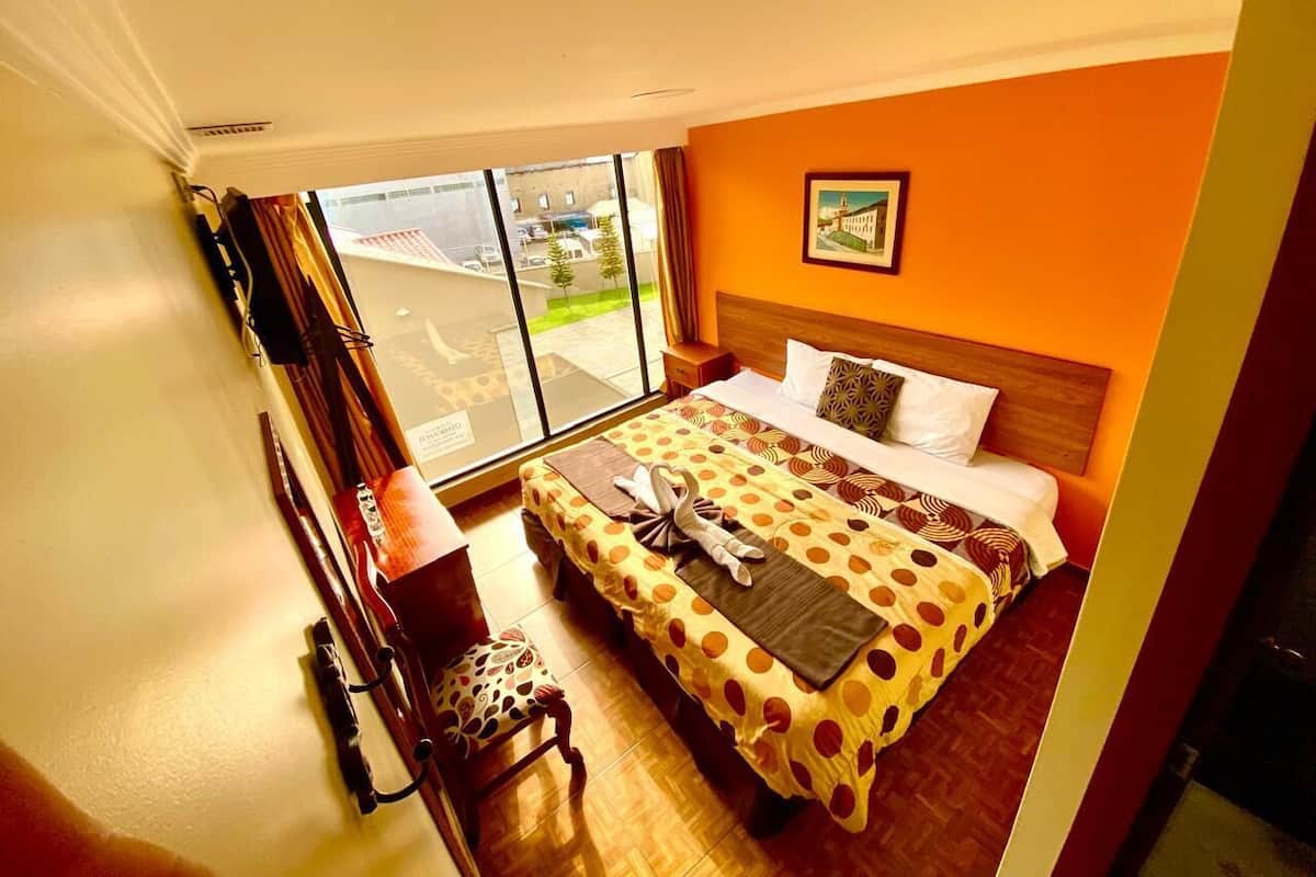 Une chambre d’hôtel avec un grand lit, une tête de lit en bois, une fenêtre donnant sur l’extérieur et une petite table avec une chaise.