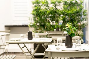 Un área de comedor al aire libre con mesas dispuestas para dos, cada una con un jarrón de flores, vasos y cubiertos.