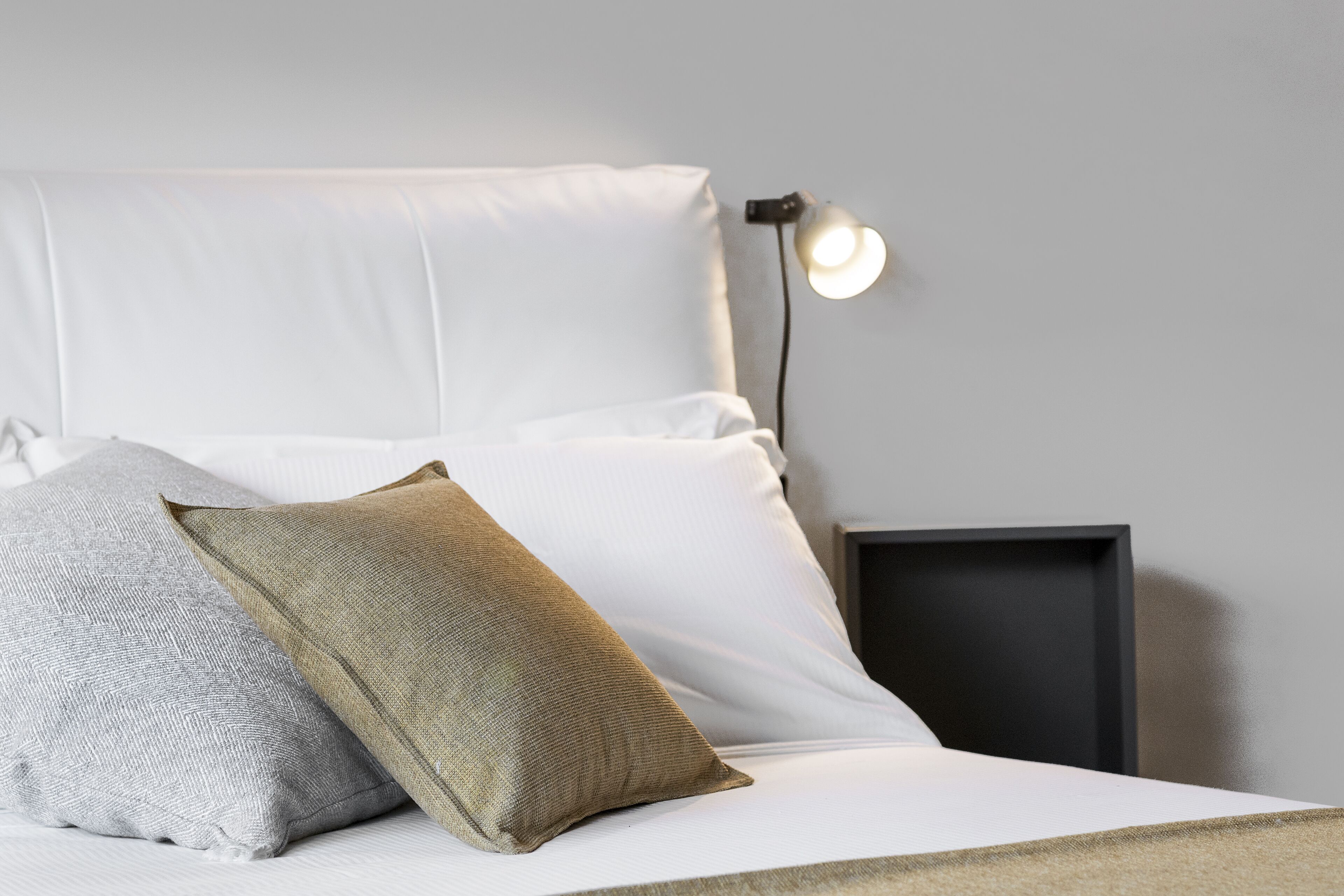 A neatly made bed with white and beige pillows, a bedside lamp, and a dark bedside table.