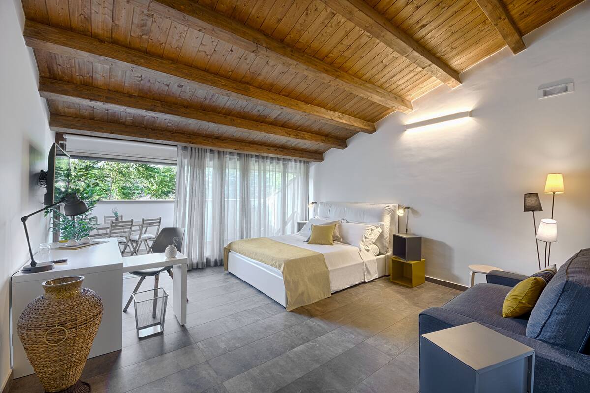 Ein Schlafzimmer mit Bett, Nachttischen, Schreibtisch, Stuhl, Sofa und einem großen Fenster mit Vorhängen.