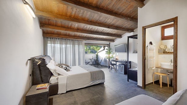 Een slaapkamer met een bed, nachtkastje en een bureau met een stoel. Er is een badkamer met een wastafel en spiegel. De kamer heeft houten balken in het plafond en een raam met gordijnen.