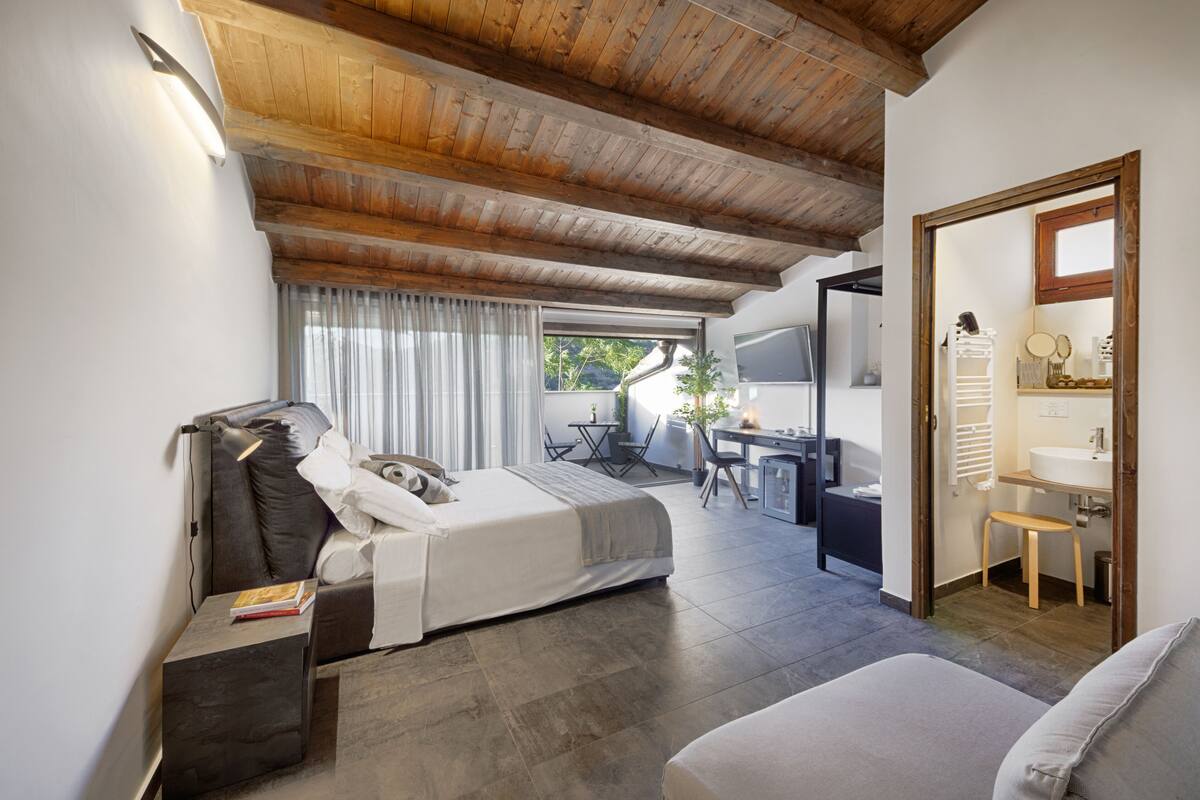 Ein Schlafzimmer mit Bett, Nachttisch und Schreibtisch mit Stuhl. Es gibt ein Badezimmer mit Waschbecken und Spiegel. Der Raum verfügt über Holzbalken an der Decke und ein Fenster mit Vorhängen.
