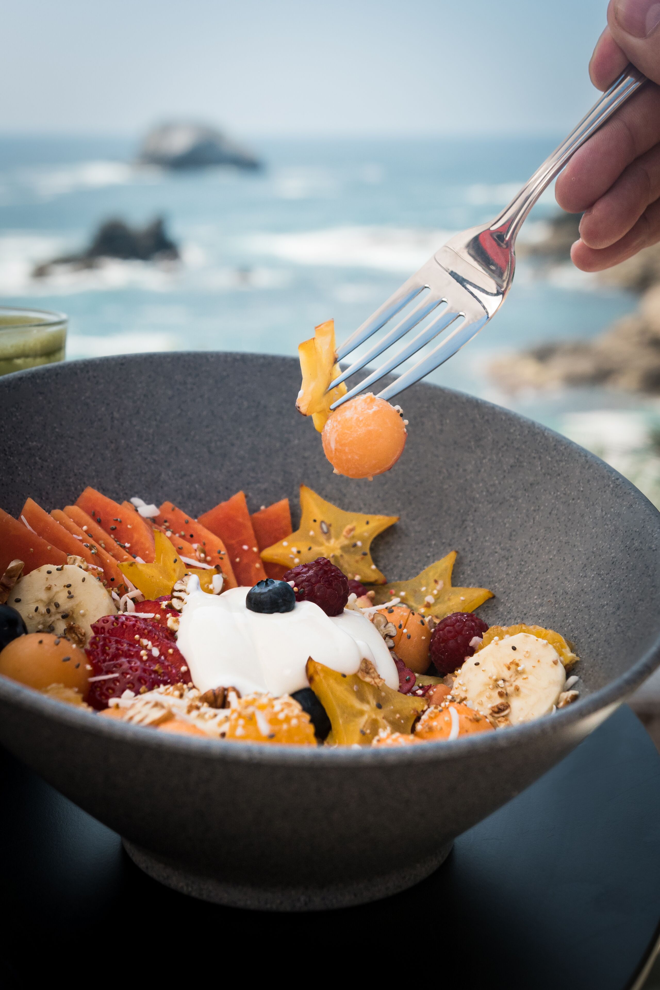 Un bol de salade de fruits, avec une fourchette qui en prend une portion, le tout présenté sur fond d’un paysage marin flou.