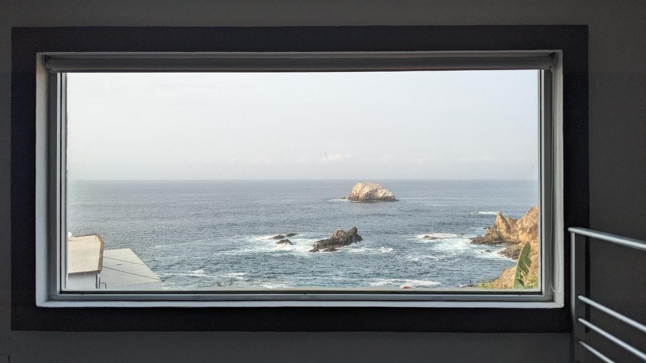 A window with a view of the ocean, a rocky outcrop, and a distant island.