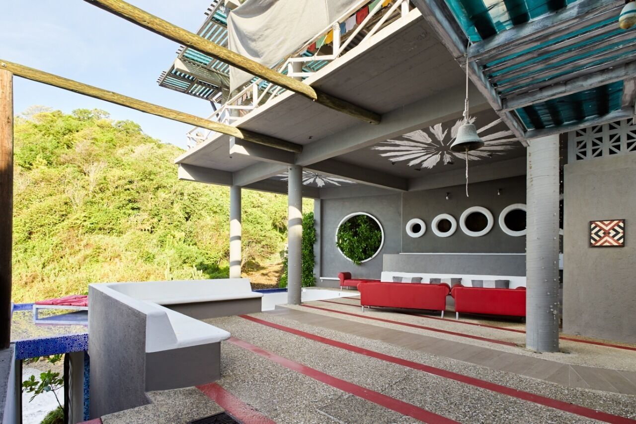 Un área de estar al aire libre con diseño moderno, que incluye un sofá rojo y sillas blancas, rodeada por una estructura de concreto con vistas a una verde ladera.