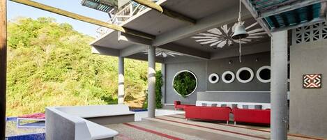 Un área de estar al aire libre con diseño moderno, que incluye un sofá rojo y sillas blancas, rodeada por una estructura de concreto con vistas a una verde ladera.