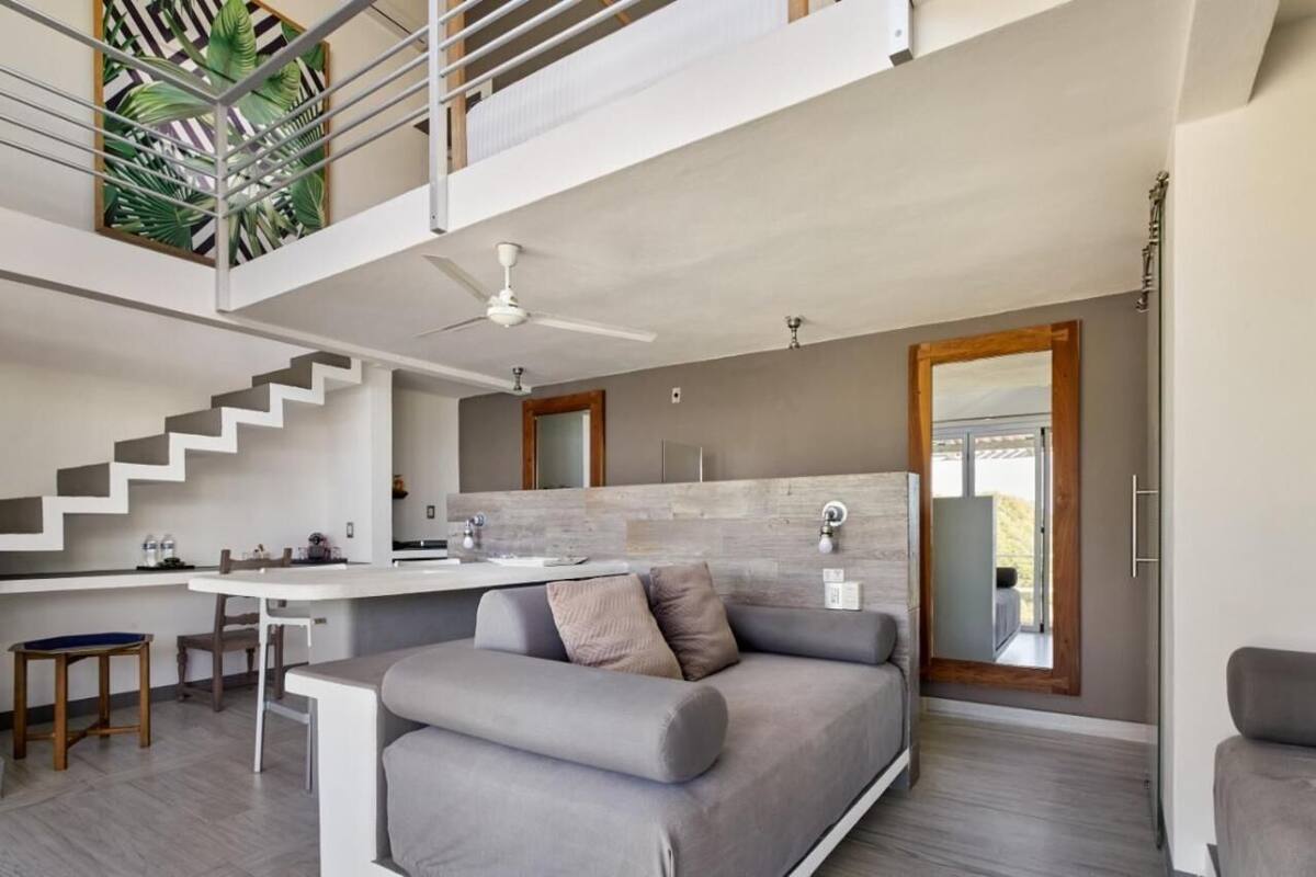 A modern interior with a staircase leading to a balcony, a kitchen area, and a seating area with a sofa and a dining table.