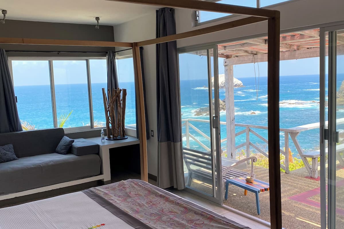 A bedroom with a large window overlooking the ocean, a sofa, a bed, and a view of the sea through a glass door.