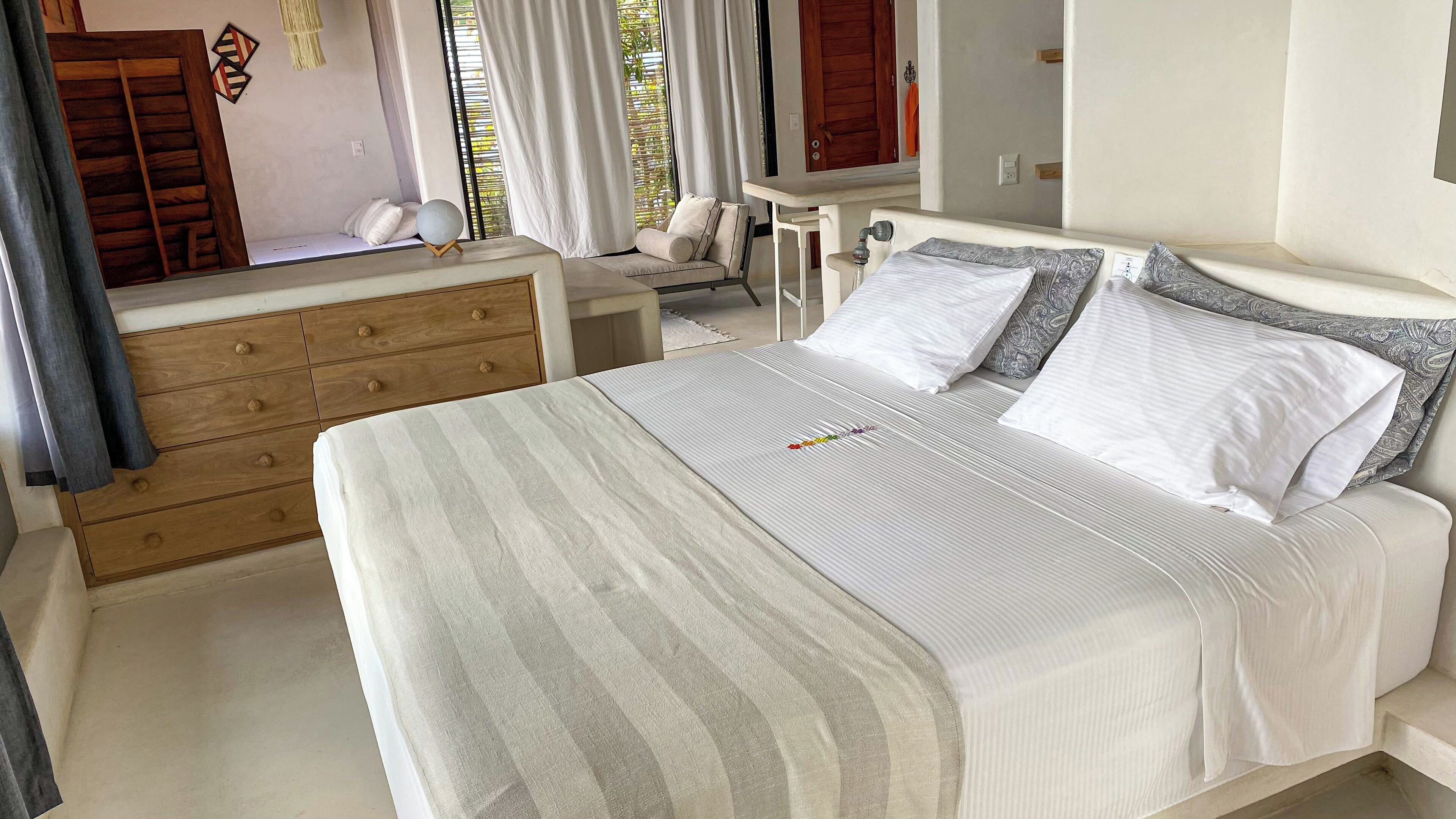 Una habitación de hotel con una cama, un aparador de madera, una mesita pequeña y una ventana con cortinas.