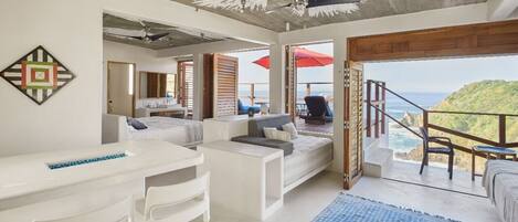 A modern bedroom with a bed, a desk, and a chair. There is a balcony with a view of the ocean.