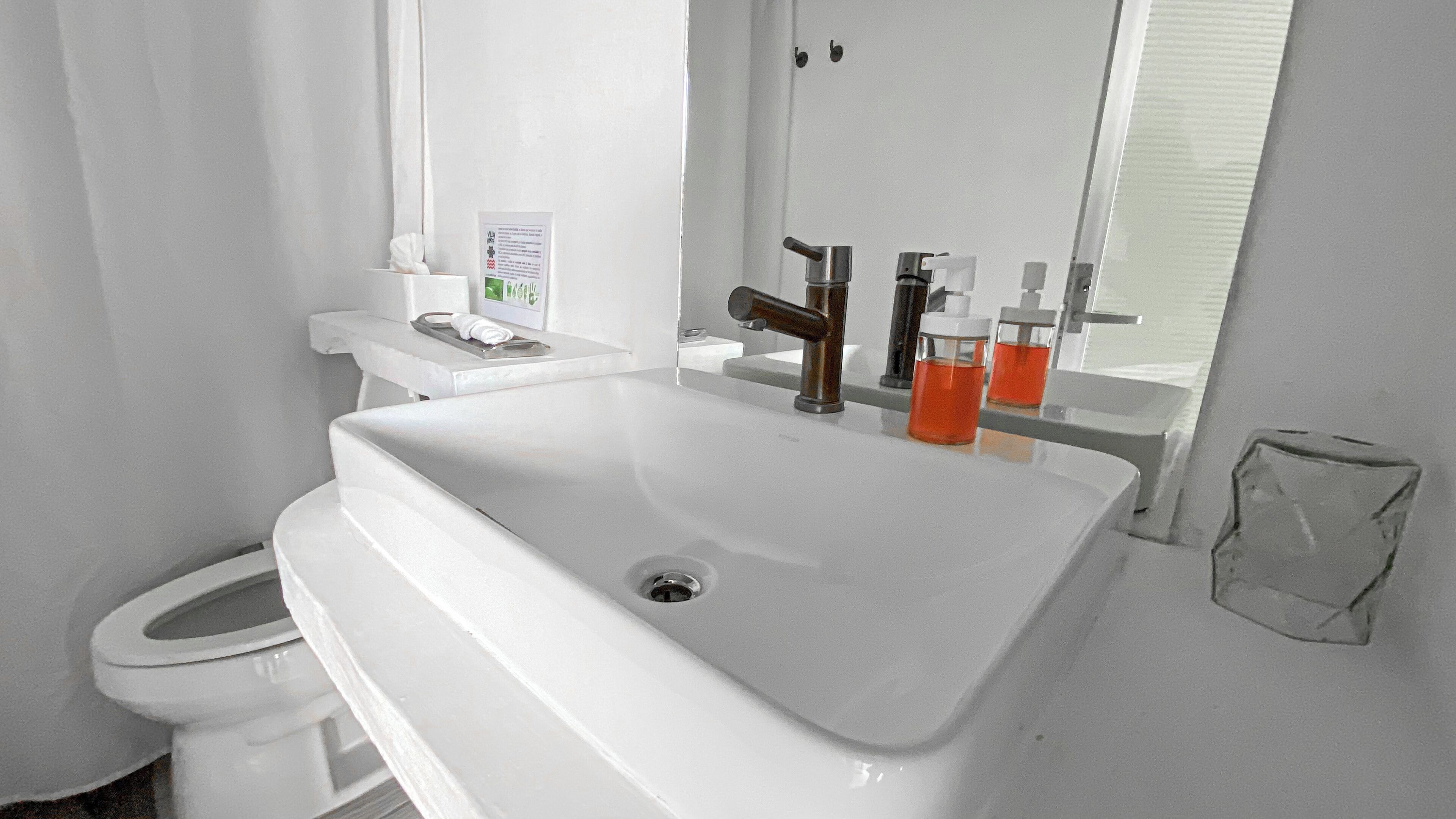 A white bathroom with a sink, a mirror, and a toilet. There are two bottles of liquid soap and a tissue box on the sink counter.