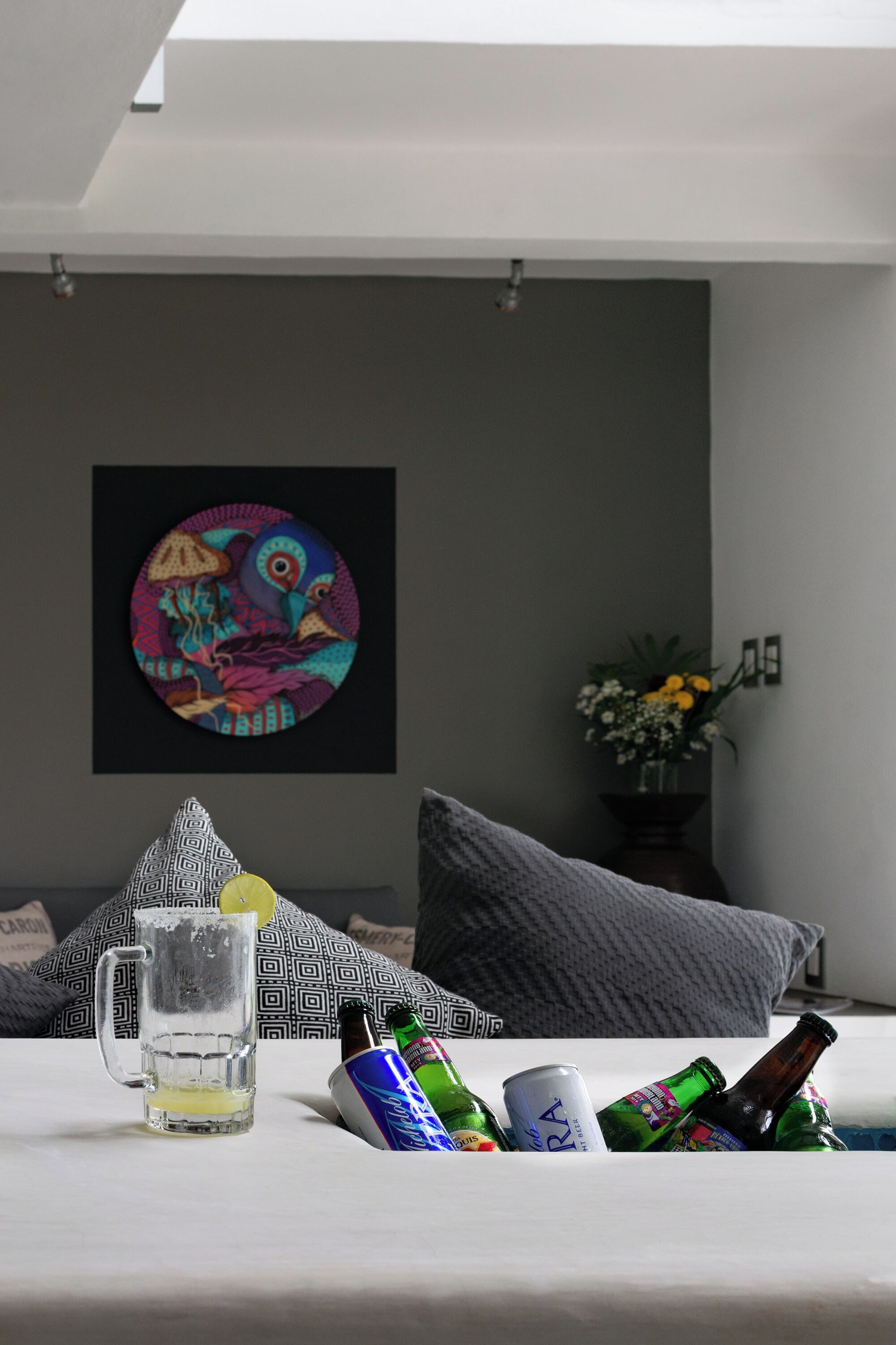 A room with a bed, a wall-mounted artwork, a vase with flowers, and a table with a mug and beer bottles.