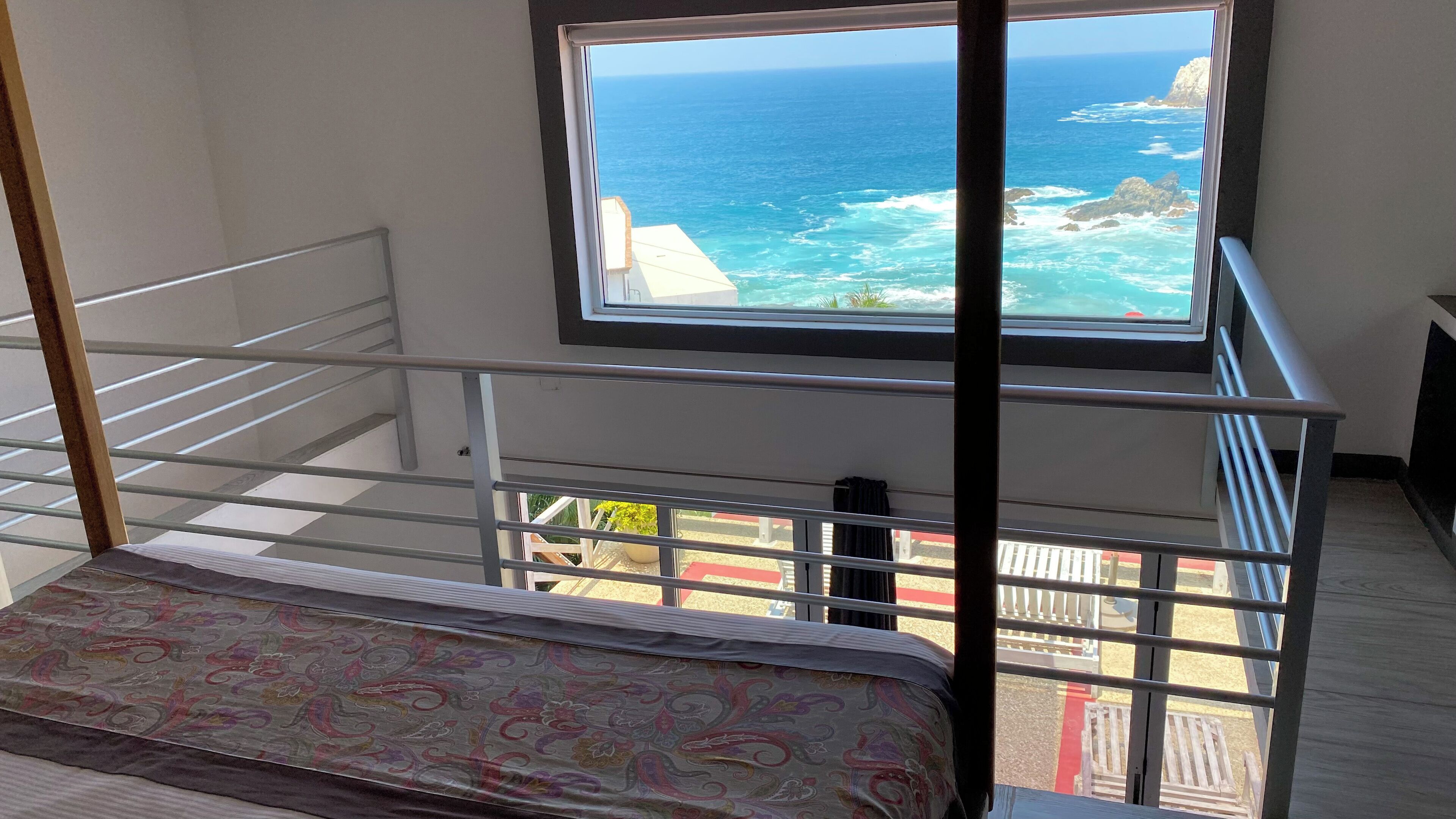 A room with a bed, a window with a view of the ocean, and a staircase.