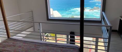 A room with a bed, a window with a view of the ocean, and a staircase.