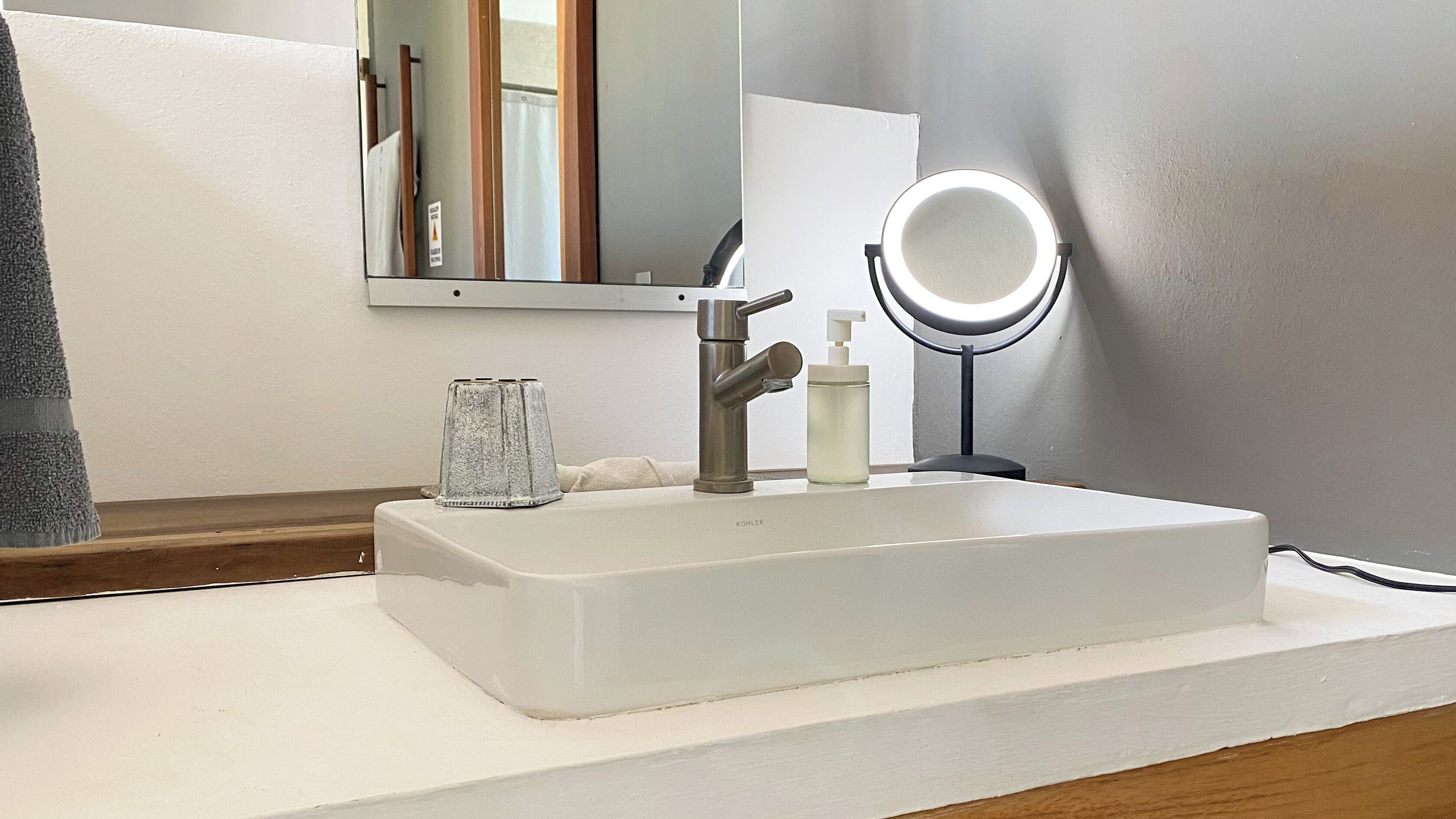 Une salle de bain avec un lavabo blanc, un robinet moderne, un récipient en verre, un distributeur de savon et un miroir rond avec un éclairage intégré.