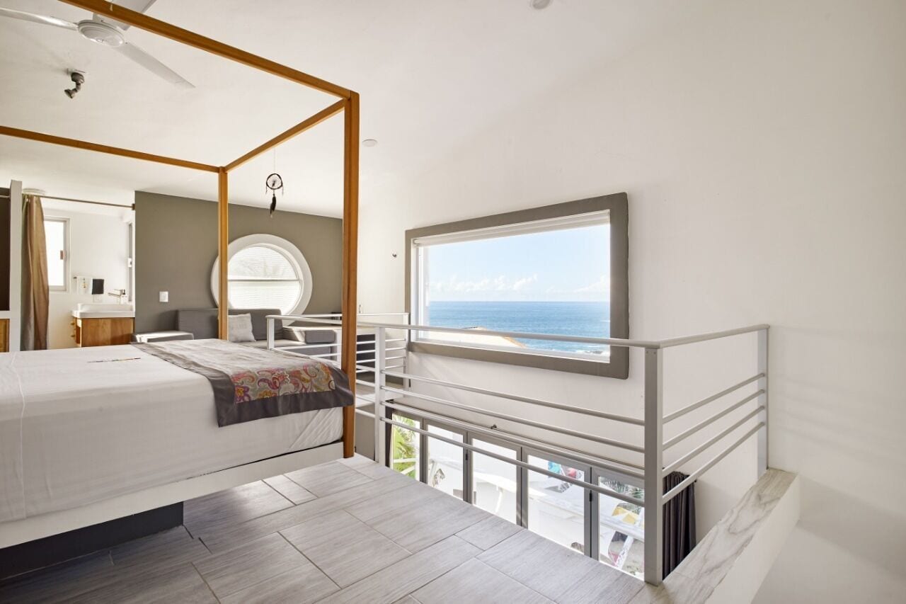 Un dormitorio con cama con dosel, un balcón con barandilla, un ventanal con vista al mar y un espejo redondo en la pared.