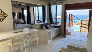 A modern room with a large window, a sofa, a small table, and a staircase leading to a balcony with a view of the ocean.