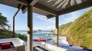 A covered outdoor seating area with steps leading to a pool, overlooking a coastal landscape with cliffs and the ocean.