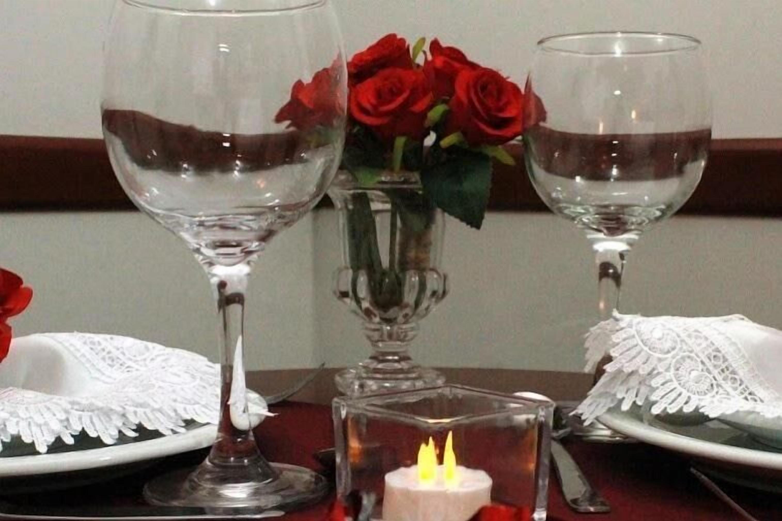 A table set with two wine glasses, a vase with red roses, a lit candle, and white lace tablecloths.
