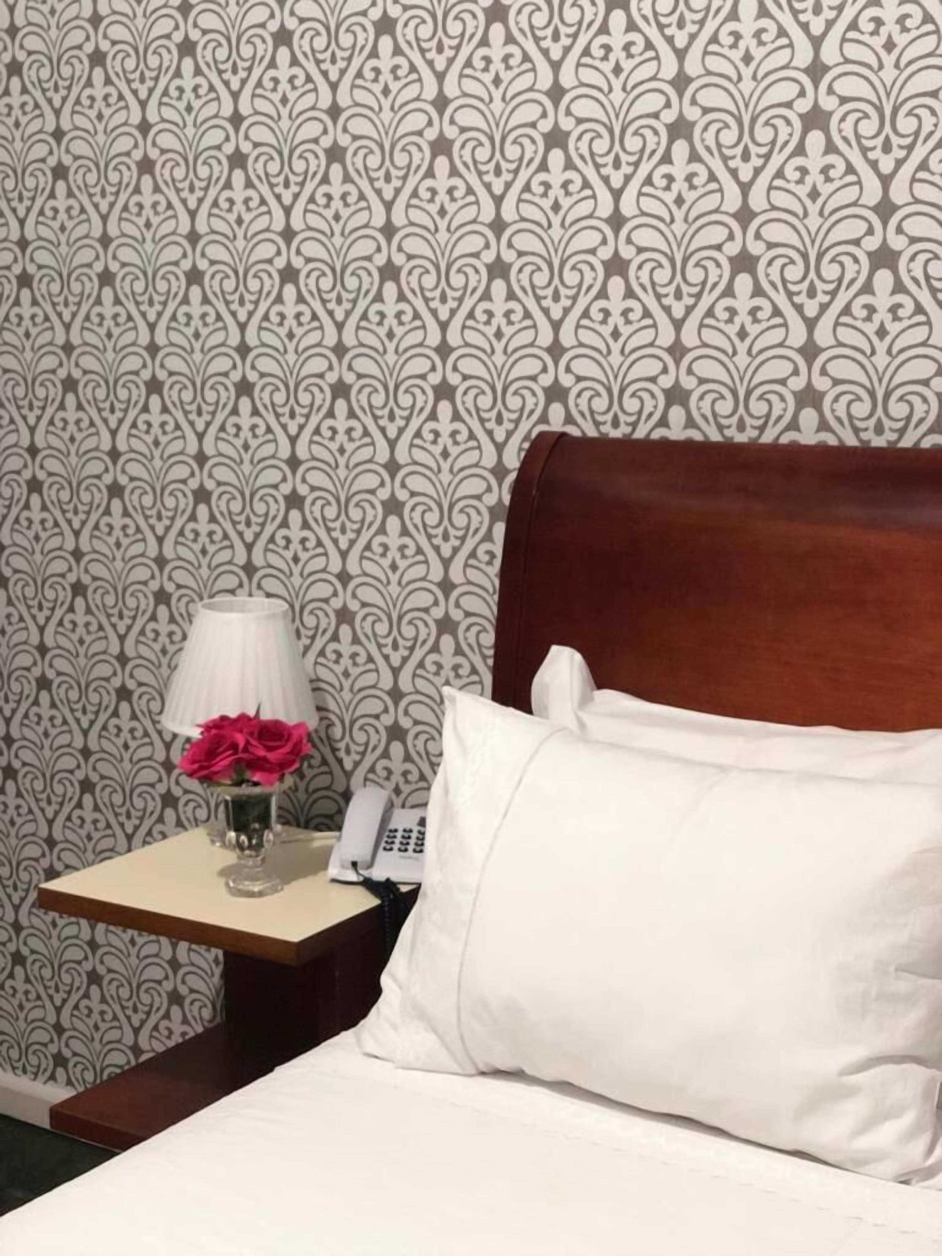 A neatly made bed with white linens and a wooden headboard, a bedside table with a lamp and a vase of flowers, and a patterned wallpaper in the background.