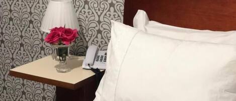 A neatly made bed with white linens and a wooden headboard, a bedside table with a lamp and a vase of flowers, and a patterned wallpaper in the background.
