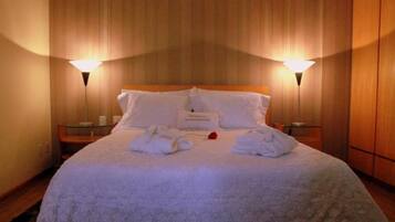 Cama bem arrumada com lençóis brancos, duas luminárias de cabeceira e uma mesa de vidro com uma vela.