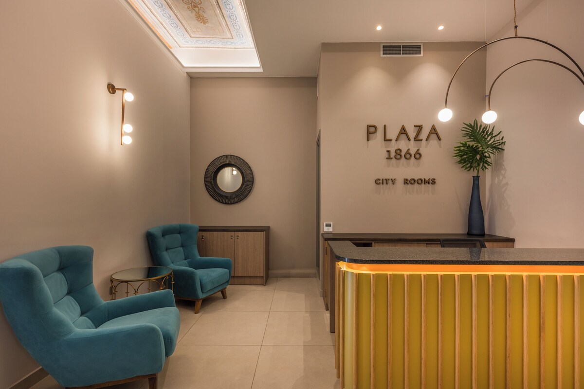 A reception area with teal chairs, a reception desk, a mirror, and a wall sign reading "PLAZA 1866 CITY ROOMS".
