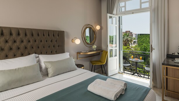 Een hotelkamer met een gestoffeerd hoofdeinde, een bed met witte lakens, een groene plaid, een klein bureau met een spiegel en een balkon met een tafel en stoelen.