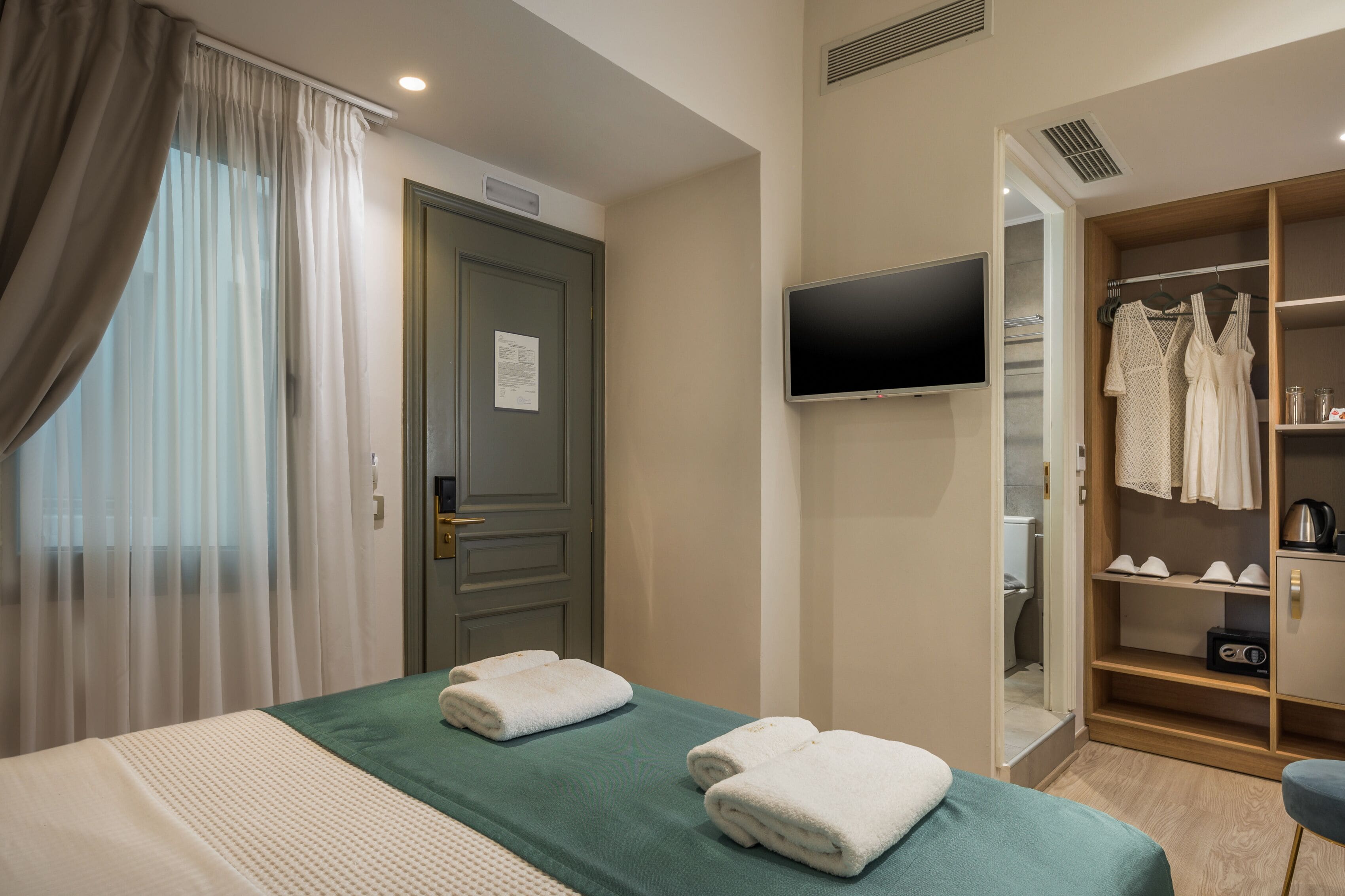 Une chambre d’hôtel avec un lit, une télévision, une armoire avec des vêtements et une fenêtre avec des rideaux.