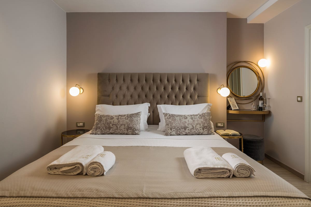 Un lit bien fait, avec deux oreillers, une table de chevet sur laquelle se trouve une lampe, et un miroir rond accroché au mur.