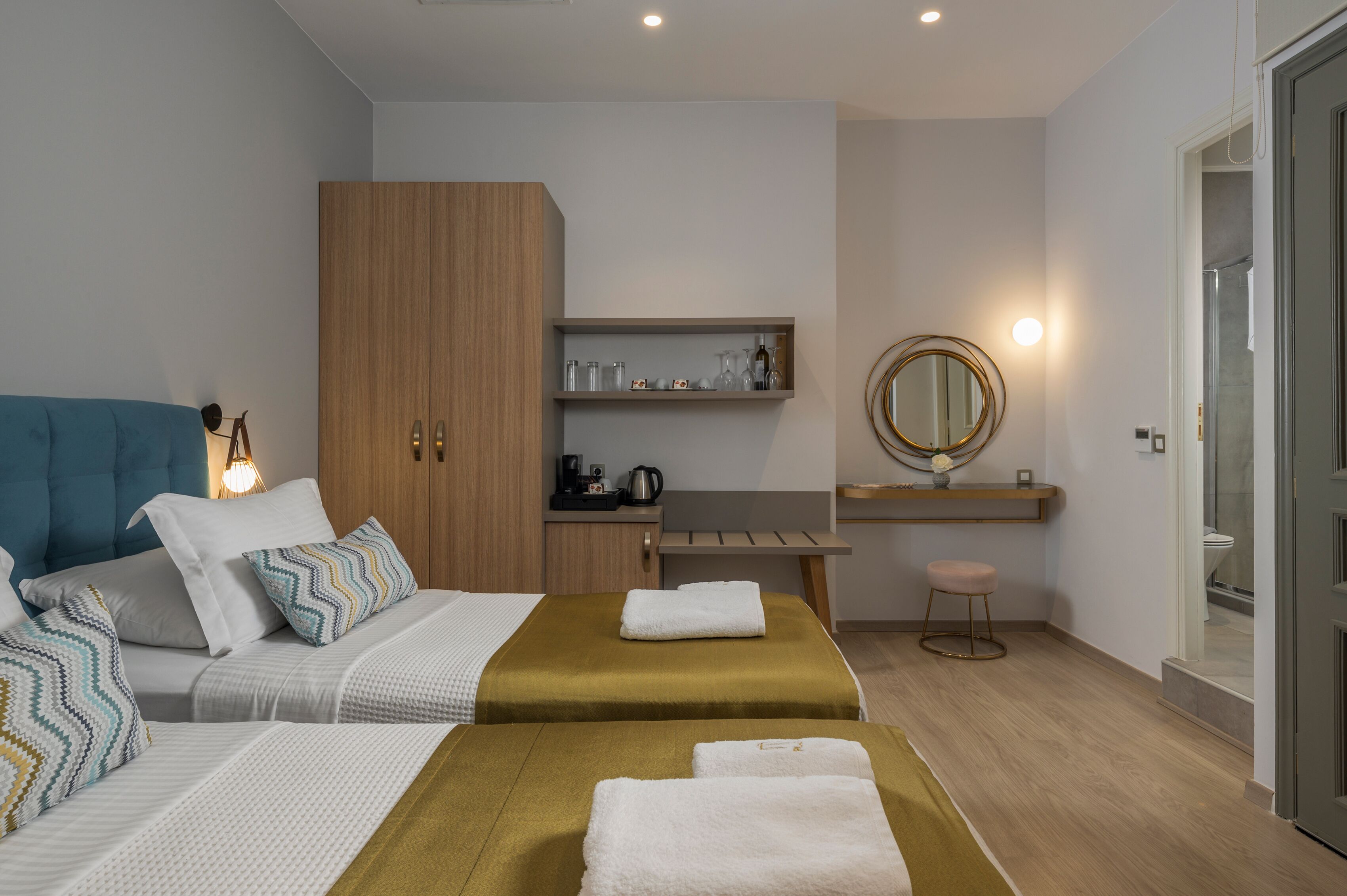 A modern hotel room with two beds, a wooden wardrobe, a small kitchenette, a bathroom with a glass shower, and a round mirror.