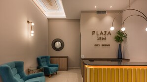 Een ontvangsthal met smaragdgroene stoelen, een receptiebalie, een spiegel en een bord met de tekst 'PLAZA 1866 CITY ROOMS'.