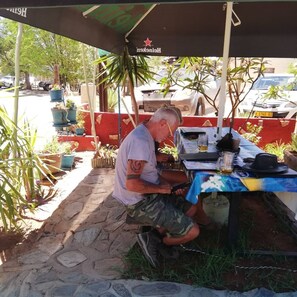 A man working at an outdoor table with a beer can, surrounded by potted plants and a red wall. - The Farmhouse B&B Beergarden CC (Outjo)