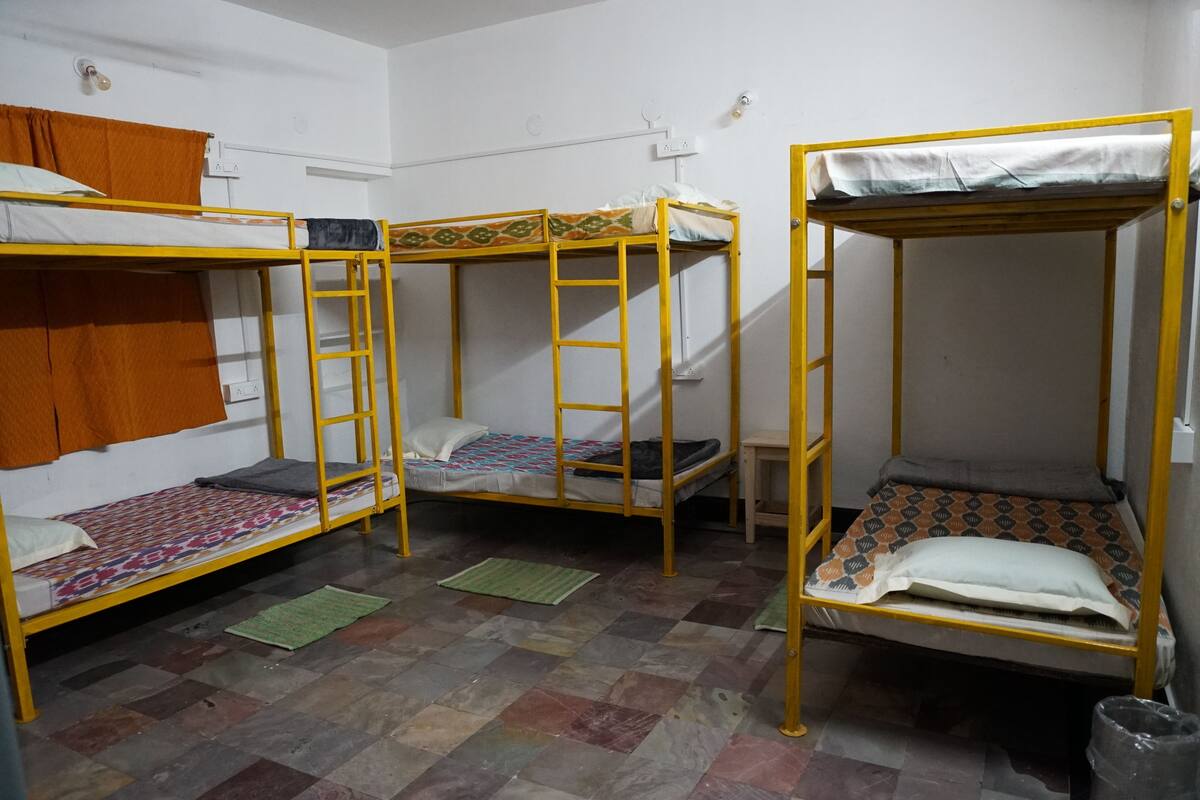 A dormitory room with three bunk beds, each with a mattress and pillow. There is a small table and a green mat on the floor.