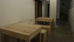 A narrow hallway with wooden tables and benches, a white wall, and a brick wall on one side. - Shepherd Stories - Hostel (Hyderabad)
