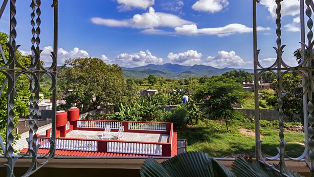 A balcony with a red railing, a white balustrade, and a view of mountains and greenery. - Hostal MJ (Trinidad)