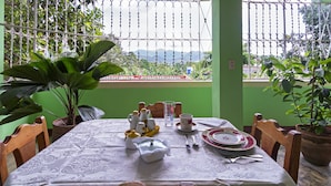 A dining table set with plates, cups, and cutlery, surrounded by chairs, with a view of mountains through a window with a metal fence. - Hostal MJ (Trinidad)