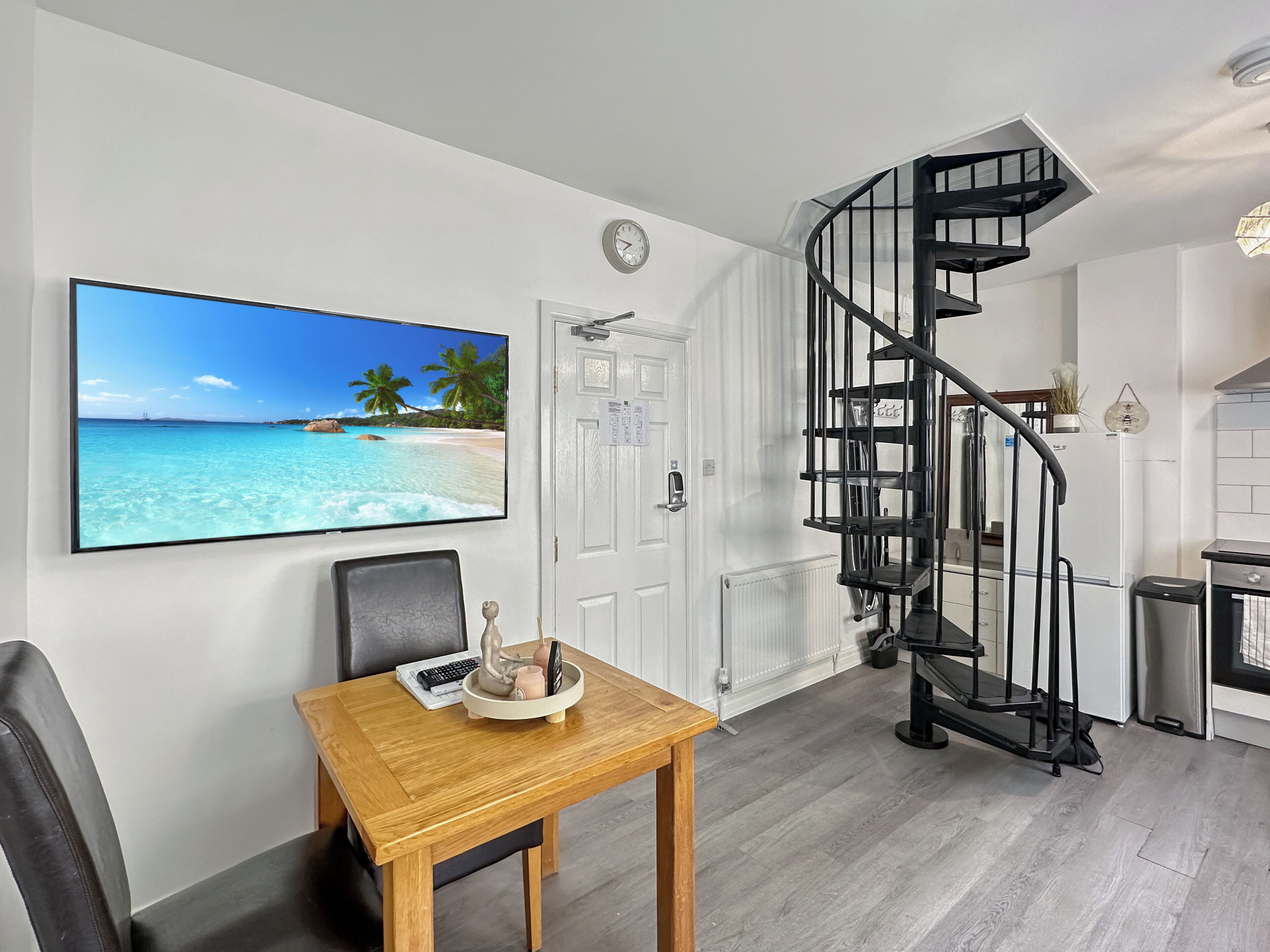 A modern kitchen with a spiral staircase, a dining area with a table and chairs, and a large beach-themed TV on the wall.