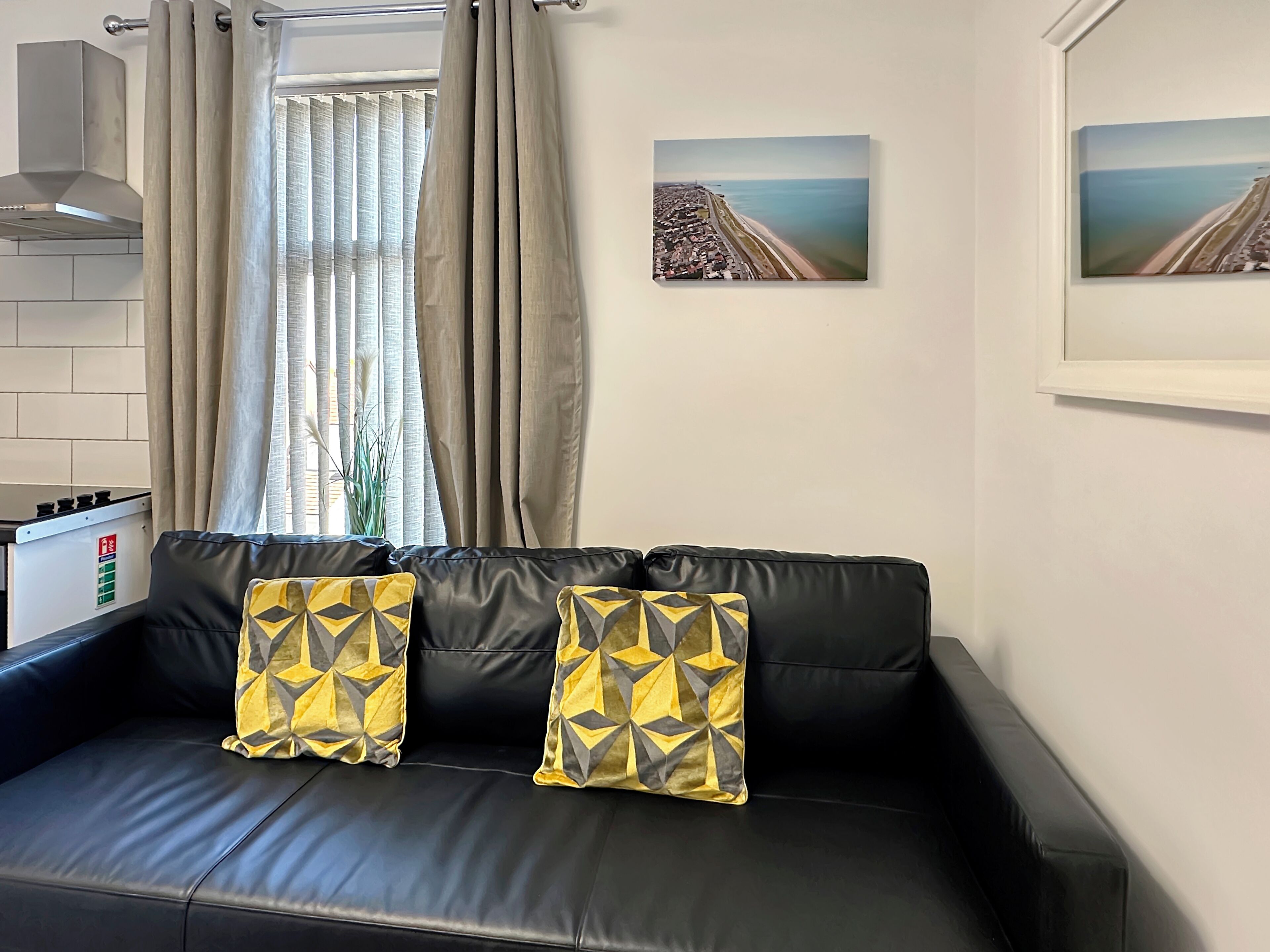 A modern living room with a black leather sofa, two patterned cushions, a window with curtains, a kitchen area, and two framed pictures on the wall.