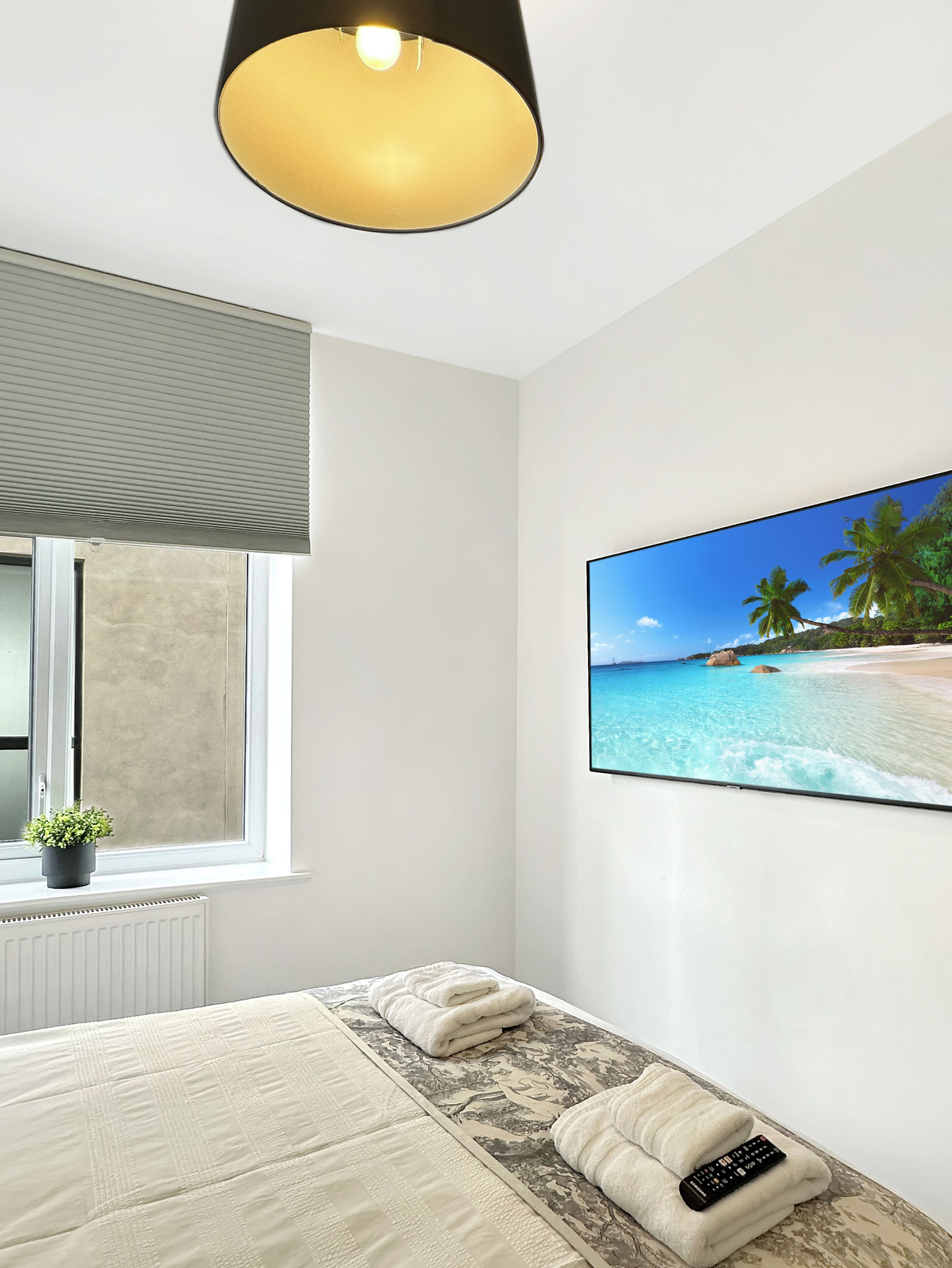 Un dormitorio con una cama, una ventana con persianas, una planta en maceta y un cuadro de una escena de playa.