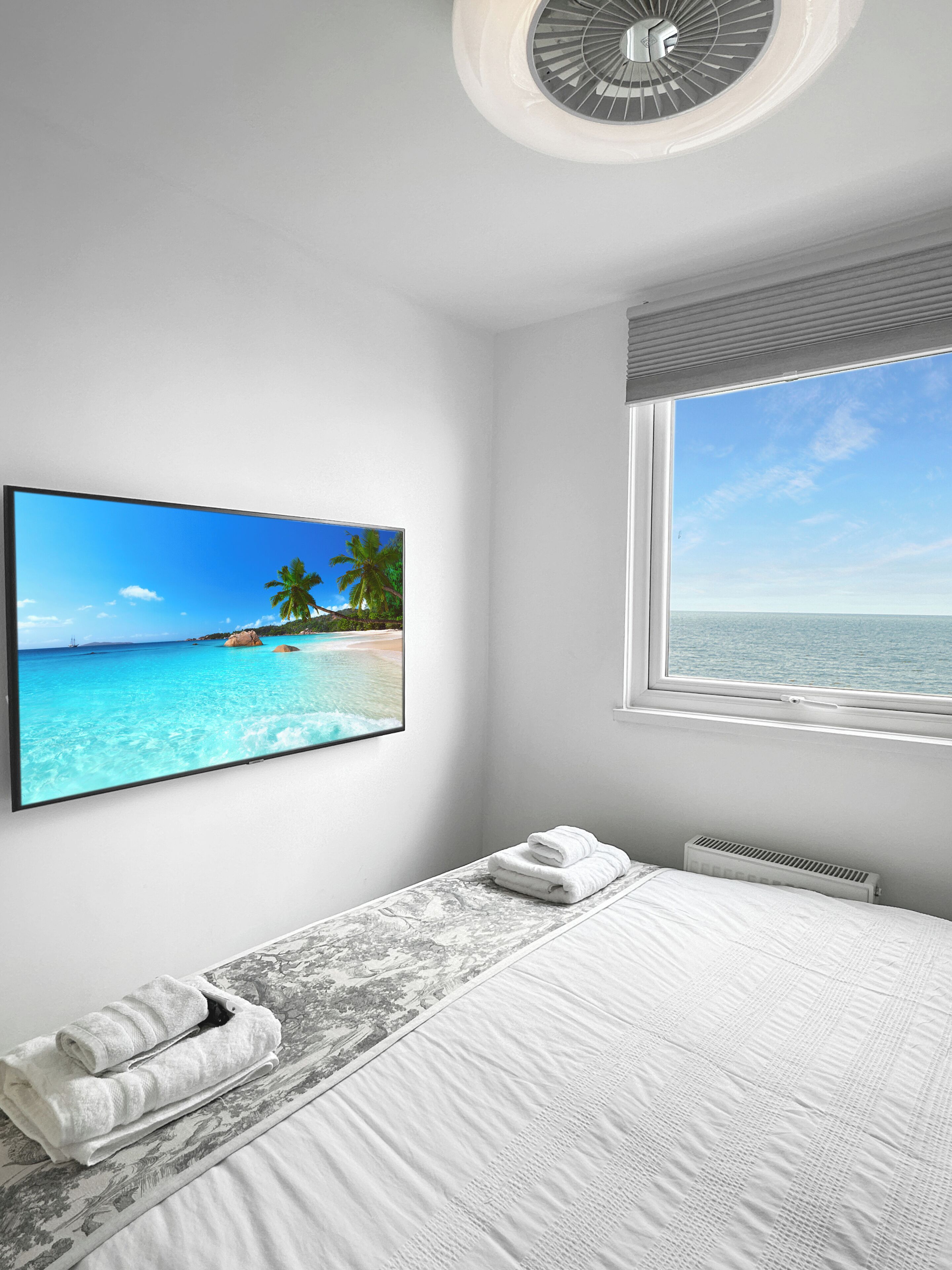 A bedroom with a bed, a window with a view of the sea, a ceiling fan, and a wall-mounted TV displaying a tropical beach scene.