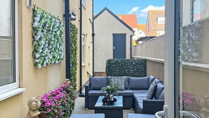 Un patio exterior estrecho con un sofá negro, dos sillas de mimbre negro y una pequeña mesa. Hay una planta decorativa con flores rosadas a la izquierda y una planta montada en la pared a la derecha.