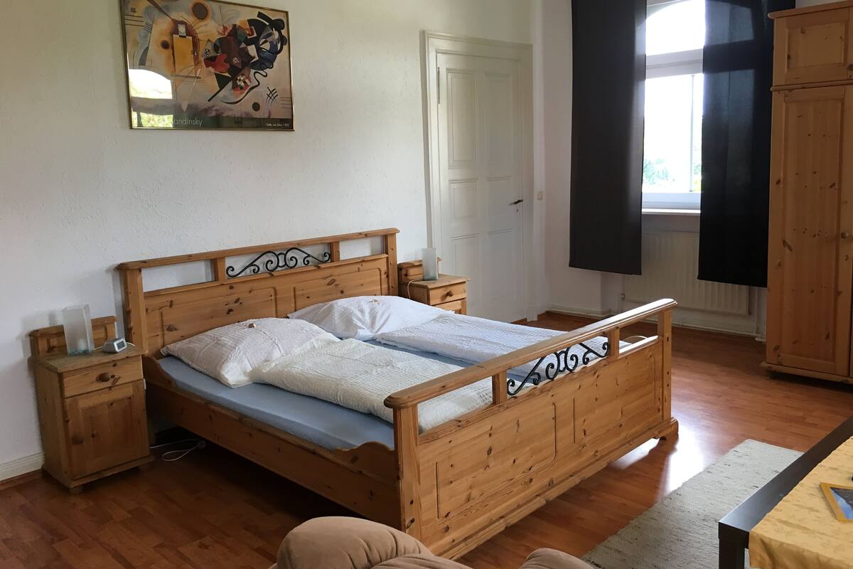 A wooden bed with a headboard, a bedside table, a nightstand, a window with black curtains, and a painting on the wall.