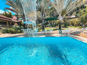 A swimming pool with clear blue water, surrounded by palm trees and outdoor furniture. - Pousada Quinta dos Colibris (São Bento do Sapucaí)