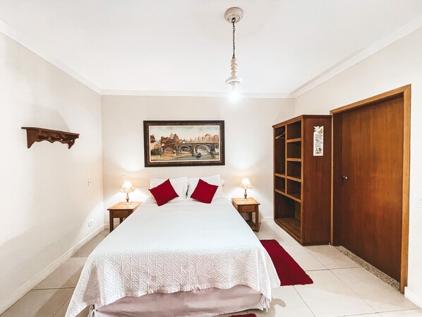 A bedroom with a bed, two bedside lamps, a wooden wardrobe, a painting on the wall, and a red rug. - Pousada Quinta dos Colibris (São Bento do Sapucaí)