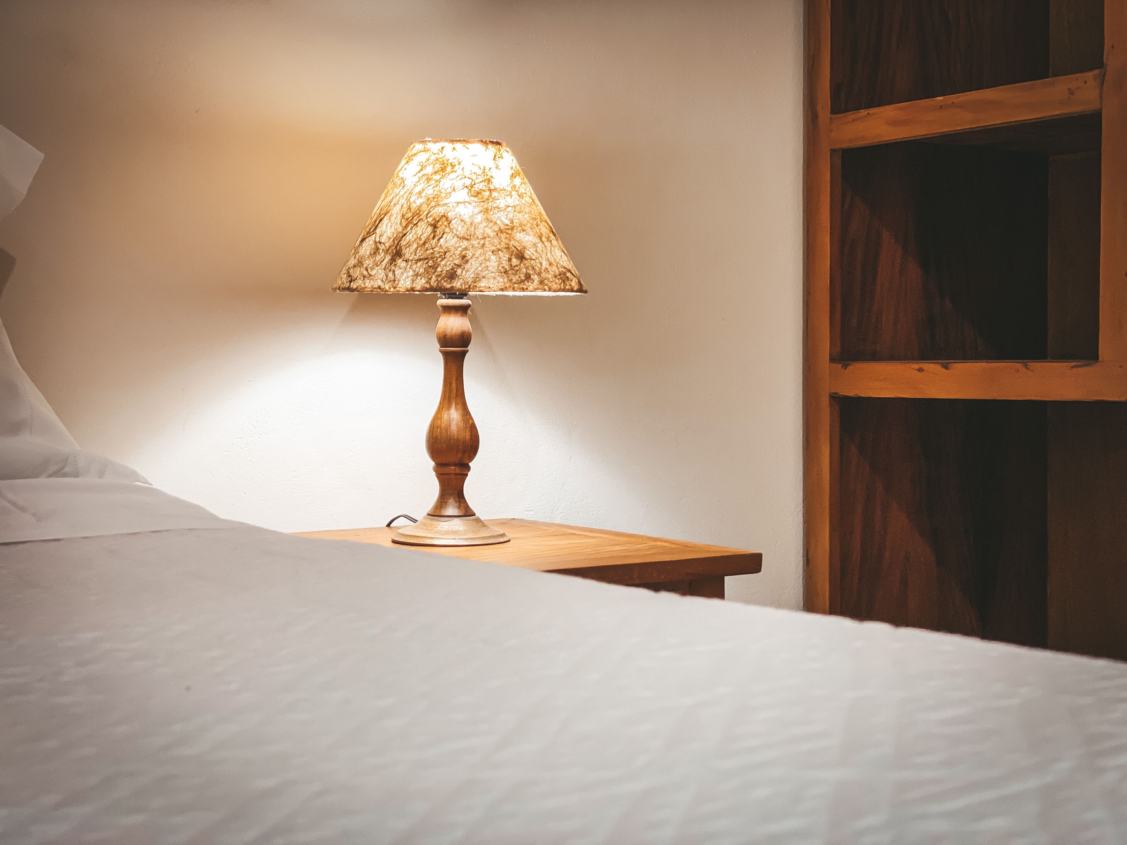 A wooden bedside table with a lamp, a bed with white linens, and a wooden wall panel.