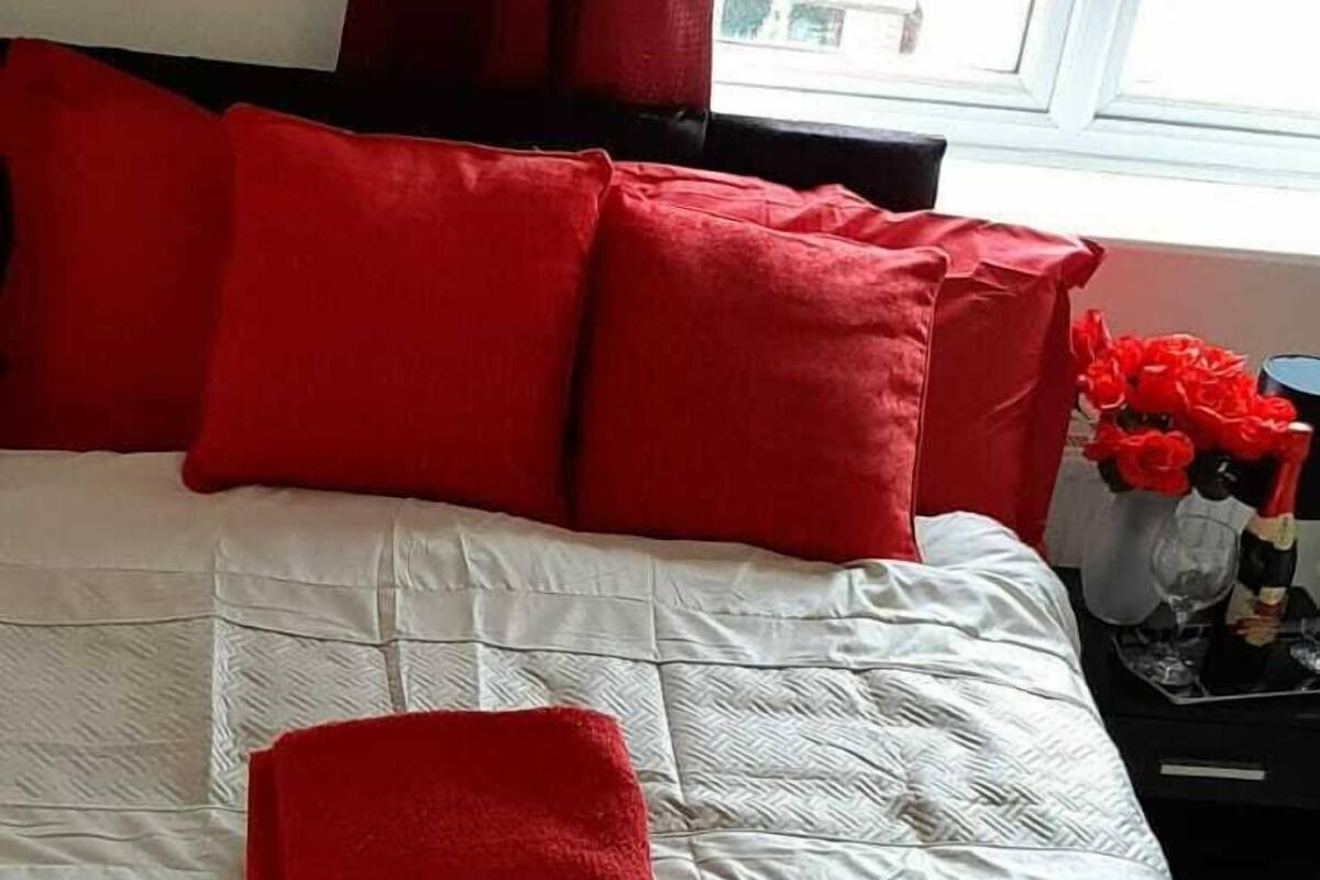 A bed with red bedding and pillows, a nightstand with a vase of red flowers, and a window with blinds.
