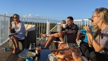 A group of people sitting on wicker chairs around a table with food and drinks, on an outdoor deck with a railing and a view of the landscape.