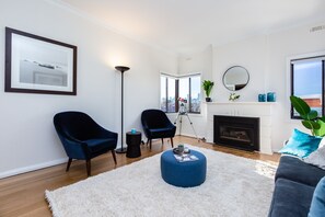 Ein modernes Wohnzimmer mit Kamin, zwei dunklen Sesseln, einem runden Hocker und einem großen Fenster mit Blick auf die Stadt.