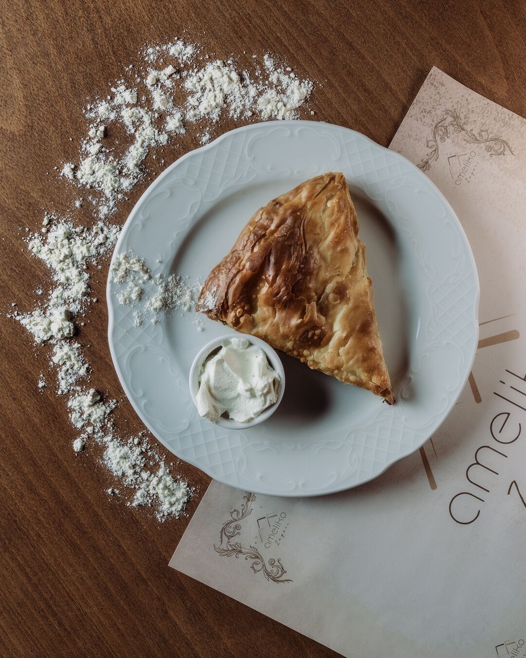 Une pâtisserie triangulaire sur une assiette, accompagnée d'un petit bol de crème, le tout entouré de miettes éparpillées sur une surface en bois.